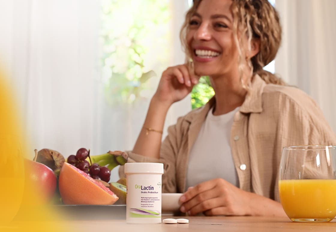 OraLactin Kaupastillen stehen auf Tisch mit Obst und Orangnsaft, im Hintergrund sitzt eine Frau OraLactin Kaupastillen stehen auf Tisch mit Obst und Orangnsaft, im Hintergrund sitzt eine Frau