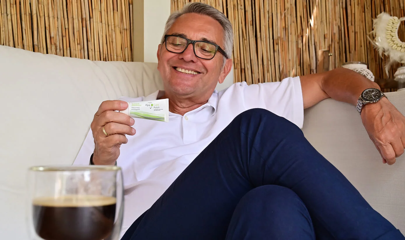 Mann hält ApaCare Zahn-Polierpaste in der Hand, vor ihm steht ein Glas mit schwarzem Kaffee Mann hält ApaCare Zahn-Polierpaste in der Hand, vor ihm steht ein Glas mit schwarzem Kaffee